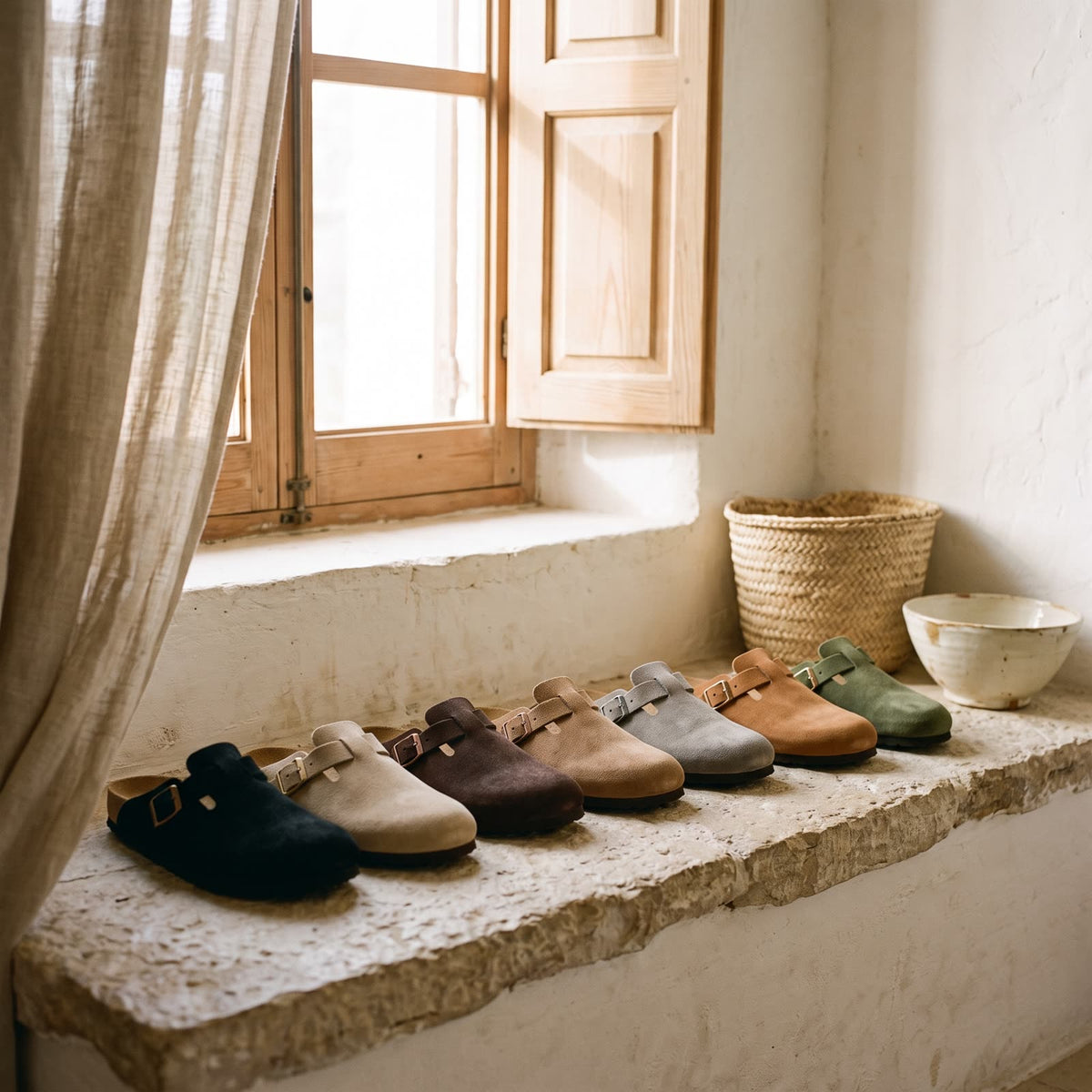 Suede clogs in black, sand, dark brown, light brown, grey, caramel and moss green on a stone ledge by a shuttered window